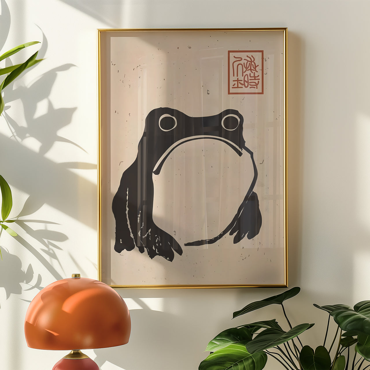Framed artwork of a black frog on beige paper with a red stamp, placed on a light wall with plants.